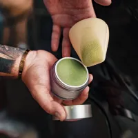 A jar of green-tinted cream is being held open, with a makeup sponge hovering above it, partially dipped into the cream. Both hands belong to a person wearing a gold bracelet.