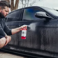 A man sprays a red cleaning solution, labeled "FÖRST" on a dirty black car door in an outdoor setting with trees in the background.