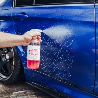 Hand sprays cleaning solution from a bottle labeled "FÖRST" onto a blue car door, which is wet and soapy. The car is parked on a wet concrete surface.