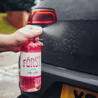 A hand sprays "FÖRST Citrus Pre-Cleaner" from a pink bottle onto a black car's rear in an outdoor setting with grass and brick nearby.