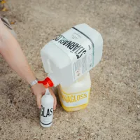 A person refills a small white bottle labeled "GLASS" from a larger white container labeled "STRAMAGLASS," placed upside down on a yellow container labeled "NAGLOSS," in an outdoor setting with a gravel ground.