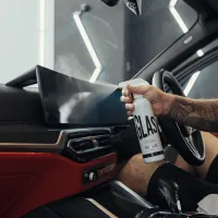 A hand sprays a car cleaner from a white bottle labeled "GLAS" onto the interior windshield of a modern car, with a red dashboard visible in a well-lit garage.