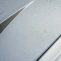 Raindrops are scattered on the silver surface of a car hood, emphasizing a sleek, metallic texture and partially separated by narrow grooves.