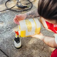 A person in red shorts pours yellow liquid from a "Stjarnagloss" container into a spray bottle on a concrete surface, with a coiled hose and soap suds nearby.