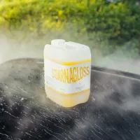 A white and yellow container of "STJÄRNAGLOSS" cleaning solution is placed on a wet, shiny surface, surrounded by mist, with a blurry green hedge in the background.