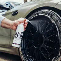 A hand sprays "HUL Colour Change Wheel Cleaner" onto a large, black car wheel coated with foam, with a green car in the background.