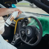 Hand sprays cleaner onto steering wheel in a modern car interior with yellow accents, using a bottle labeled "Interior Cleaner - HIGHWAY STAR CLASS." Green cloth draped over the dashboard.