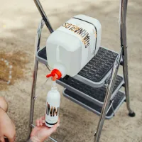 A large bottle labeled "SILIKANGLOS" dispenses liquid into a smaller bottle labeled "INN" via a red nozzle, placed on a step ladder, outdoors.