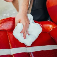 A hand holding a white cloth wipes the surface of a red leather car seat, with some cleaning product applied, inside a vehicle.