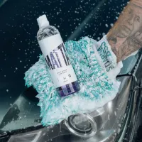 A gloved hand holding a bottle labeled "MATTE" along with "MATT FINISH CAR WASH, STJARNAGLOSS" on a sudsy, green microfiber mitt, beside a headlight on a wet, dark car.