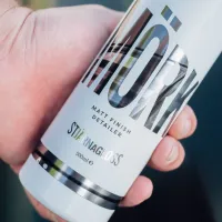 Hand holding a bottle labeled "MÖRK" with additional text "MATT FINISH DETAILER," "STJARNAGLOSS," and "500ml e" in an outdoor setting.