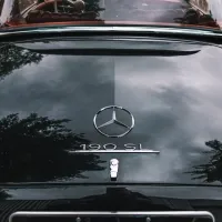 A black vintage car with a Mercedes-Benz emblem and "190 SL" text on the trunk, reflecting trees and a cloudy sky, with a partial view of the car's interior.