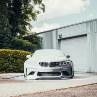 A car covered in white foam is parked in front of a large, industrial metal building with a number "12" above a closed garage door; greenery is on the left.