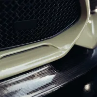 The close-up photograph shows the front grille of a car, with a cream-colored bumper and a carbon fiber lower lip detail, under low lighting conditions.