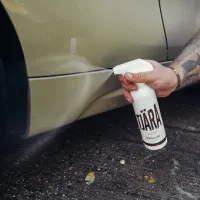 A tattooed hand sprays Tjäragloss Tjära onto a beige car's lower panel, with pavement and small debris scattered around. Text on the spray bottle reads: “TJÄRA, Tar and Glue, Stjärnagloss.”
