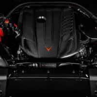 A carbon fiber engine block, adorned with a red V-shaped emblem, is situated within a sleek vehicle hood, highlighting red accents and various mechanical components in the surrounding environment.