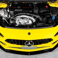 A Mercedes-AMG engine is mounted in a bright yellow car with its hood open, against a concrete floor backdrop. The engine has "AMG" and the Mercedes logo on the engine cover.