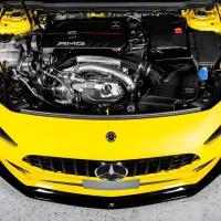 Engine compartment of a yellow car with "AMG" branding on the engine cover, positioned in a well-lit garage setting. The hood is open, revealing various engine components and details.