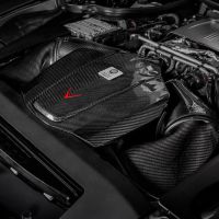 An engine with carbon fiber components situated in a sleek, modern engine bay. The carbon fiber cover features a red arrow and a label with the text "HANDCRAFTED By S. Bucem, Affalterbach."