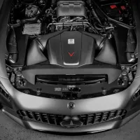A Mercedes-Benz car with its hood open exposes a detailed engine in a well-lit garage. The engine bay is meticulously organized, showcasing various components, wires, and branding details.