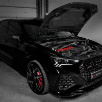 A black car with its hood open reveals a red-accented engine, parked indoors near a closed garage door. The front license plate reads "EVENTURI."