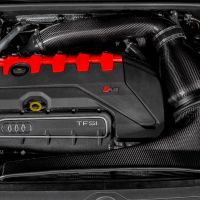 Engine bay with a prominent black and red engine cover showing "RS" and "TFSI" logos, surrounded by carbon fiber air intake pipes and electrical components in a car's compartment.