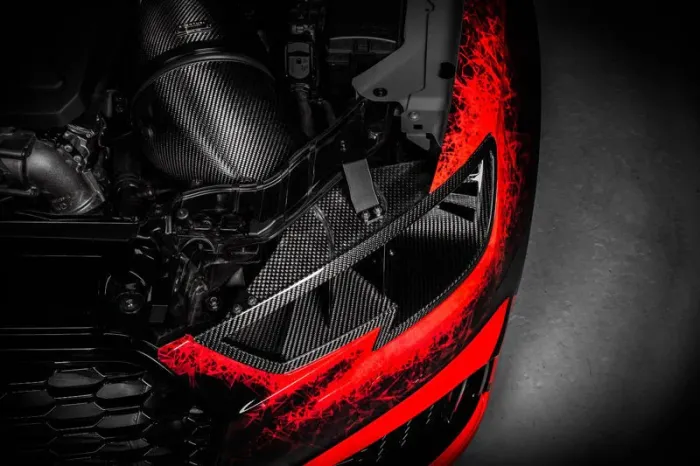 A car engine with carbon fiber components is visible under a partially removed hood in a dark environment, highlighted by striking red accents on the surrounding parts.