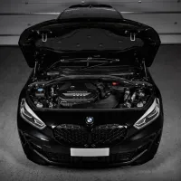 A black BMW car with its hood open, showcasing the engine, is parked in a garage with closed shutter doors in the background.
