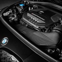 A BMW car engine marked with "M Performance" text is securely fitted in the hood, surrounded by various mechanical components. The car is parked in a dimly lit garage.