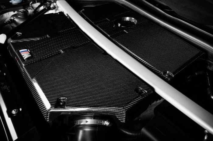 A sleek carbon fiber engine cover with a small section revealing a logo and colored stripes under the hood of a car, with visible mechanical components surrounding it.