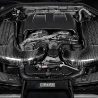A car engine equipped with polished carbon-fiber intake manifolds, located in a dark engine bay with various metal and plastic components.