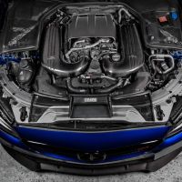 Engine with carbon components positioned within the open hood of a blue vehicle, showcasing various mechanical parts and fine details, placed on a concrete floor in a garage-like setting.