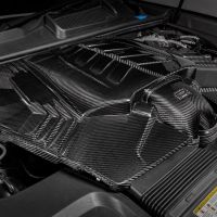 An engine compartment with a carbon fiber cover, surrounded by various mechanical components and fluid reservoirs. Text visible on the cover reads: "V12 Made in Germany." Yellow warning label at the bottom.