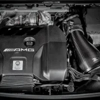 An AMG branded car engine with a carbon-fiber cover, pipes, and various components, displayed within the engine bay of a vehicle. Text: "AMG, Handcrafted by Marco Roger AMG Germany."