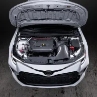 Car engine compartment with hood open, revealing various components including a carbon fiber air intake system, set in a dimly lit garage with concrete floor.