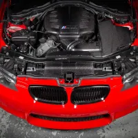 Red car hood open, exposing a complex engine with carbon fiber components, in a garage setting with a concrete floor. The engine has a logo with three colored stripes.