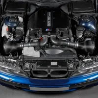 Engine displayed top-down in a blue car's hood, marked "BMW M Power." Nearby components include air filters, coolant reservoir, hoses, and electric elements, all in a garage setting.