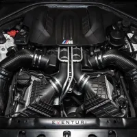 A high-performance BMW engine bay featuring carbon fiber intake components and the "M" logo, surrounded by various engine parts and housed within the car's chassis. The text "EVENTURI" is visible at the bottom.