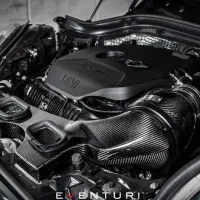 Engine covered in black casing labeled "MINI" and "Twin Power Turbo," integrated with carbon fiber intakes and piping by Eventuri, displayed in a car's open hood against a garage backdrop.