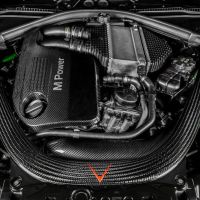 A high-performance engine with "M Power" branding is surrounded by various hoses and components, all encased in a sleek, black carbon fiber cover with bright green accents in the engine bay.