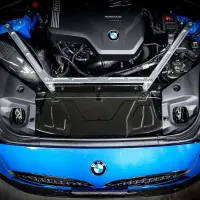 A BMW engine with "TwinPower Turbo" text displayed, mounted in the front of a blue car, secured with metallic struts and surrounding components, viewed from above in a garage setting.