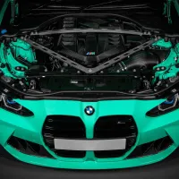 A teal BMW car with its hood open, revealing a detailed view of the engine bay, featuring a carbon fiber engine cover and structural support braces, against a dark background.