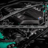 A high-performance car engine with carbon fiber components, marked with a blue, violet, and red "M" badge, encased in a green chassis, and supported by metallic struts.