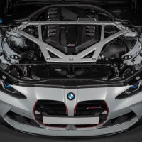 A BMW car with its hood open displays a powerful engine enclosed in a carbon-fiber cover, indicated by the BMW logo and 'M' badge, with some structural components visible.