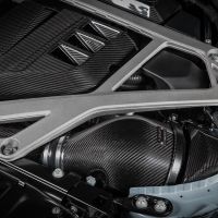 Carbon fiber engine cover with an "M" logo under a metallic strut brace; part of a high-performance vehicle's engine bay with visible wiring and components.