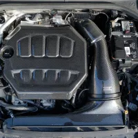 Carbon fiber engine cover and air intake system within a car engine bay, surrounded by various mechanical components, wiring, and a battery with visible labels and connectors.