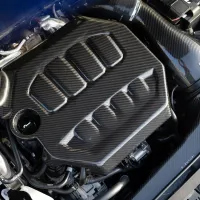 A car engine features a prominent carbon fiber cover with vent openings. The surrounding environment includes various tubes and components. The text "EVENTURI" is visible on a carbon fiber part.