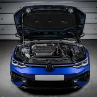 A blue car with its hood open, revealing the engine compartment in a dimly lit garage with grey floor and closed garage doors in the background.