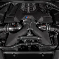 The BMW engine is situated in a car's engine bay, featuring "BMW M Power" text on the cover and surrounded by carbon fiber components and various hoses.