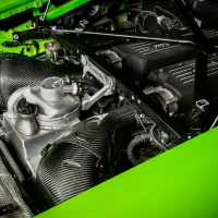 Engine compartment with "Lamborghini" on dual black air intakes, metallic components, and braided carbon-fiber pipes, surrounded by green-painted framework.